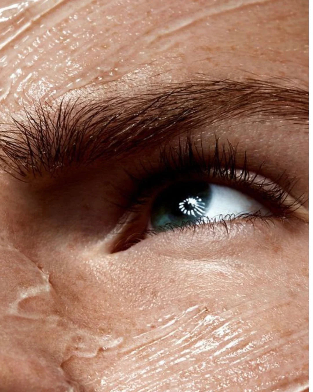 Gros plan d’un œil et d’un sourcil, avec une crème de soin brillante étalée sous l’œil.