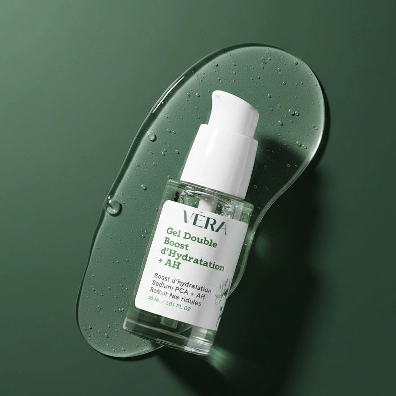 Flacon VERA Gel Double Boost d’Hydratation + AH incliné sur une goutte de gel translucide avec bulles, sur fond vert