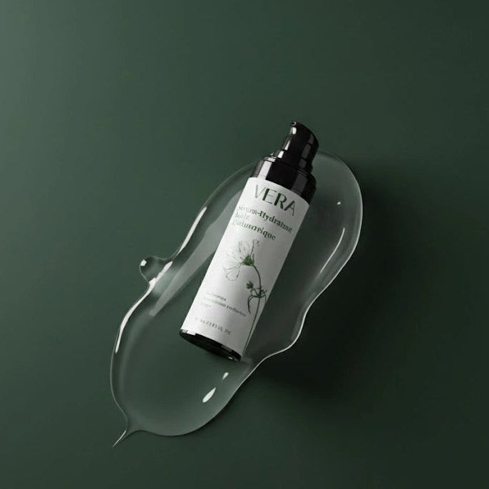 Flacon VERA du sérum hydratant à l’acide hyaluronique, en diagonale dans une goutte de gel transparent sur fond vert.